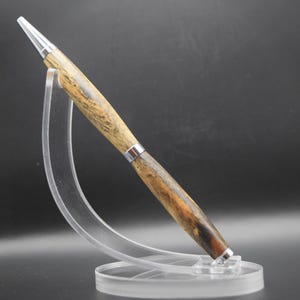 May include: A handcrafted wooden pen with a silver-colored tip and accents. The pen features a natural wood grain pattern in shades of brown and tan. It is displayed on a clear acrylic stand, showcasing its elegant design.