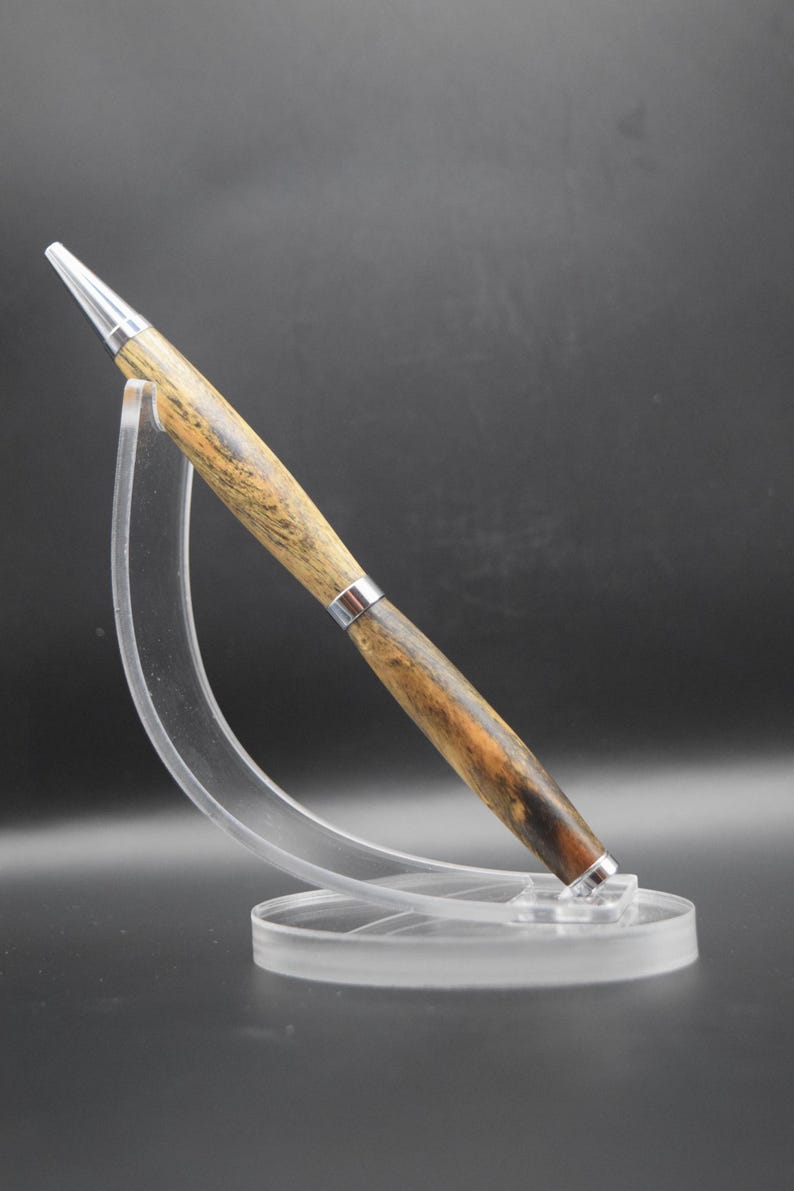 May include: A handcrafted wooden pen with a silver-colored tip and accents. The pen features a natural wood grain pattern in shades of brown and tan. It is displayed on a clear acrylic stand, showcasing its elegant design.