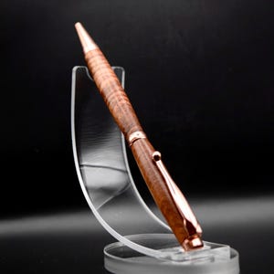 May include: A handcrafted pen with a brown wooden barrel and rose gold accents. The pen is displayed on a clear acrylic stand against a black background. The pen's design features a textured wood grain pattern.