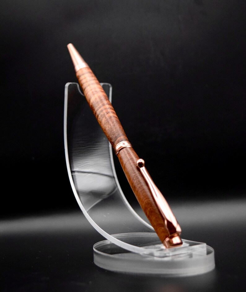 May include: A handcrafted pen with a brown wooden barrel and rose gold accents. The pen is displayed on a clear acrylic stand against a black background. The pen's design features a textured wood grain pattern.