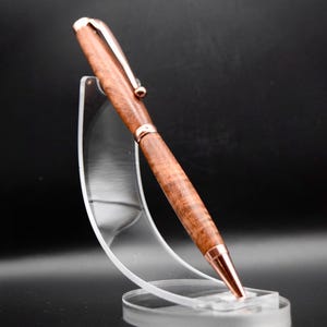 May include: A handcrafted wooden pen with a rose gold-colored clip, tip, and accents. The pen is displayed on a clear acrylic stand against a dark background. The wood grain is visible, showcasing a natural, warm brown color.