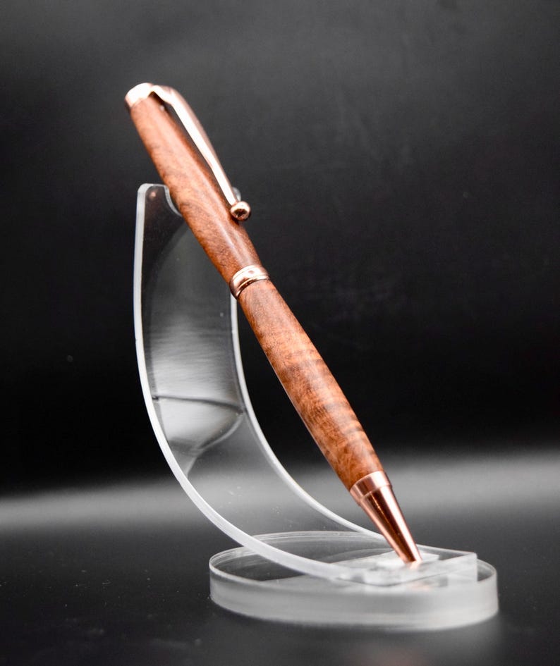May include: A handcrafted wooden pen with a rose gold-colored clip, tip, and accents. The pen is displayed on a clear acrylic stand against a dark background. The wood grain is visible, showcasing a natural, warm brown color.