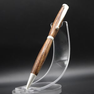Könnte beinhalten: Ein handgefertigter Holzstift mit silberfarbenen Akzenten. Der Stift hat einen dunkelbraunen Holzschaft und eine silberne Spitze und einen Clip. Er wird auf einem transparenten Acrylständer vor schwarzem Hintergrund präsentiert.