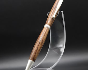 Puede incluir: Un bolígrafo de madera artesanal con detalles plateados. El bolígrafo presenta un cuerpo de madera de color marrón oscuro y una punta y clip plateados. Se exhibe sobre un soporte de acrílico transparente sobre un fondo negro.