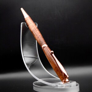 May include: A handcrafted wooden pen with a copper-colored clip and tip. The pen features a detailed wood grain pattern and is displayed on a clear acrylic stand against a dark background. The pen is a stylish writing instrument.