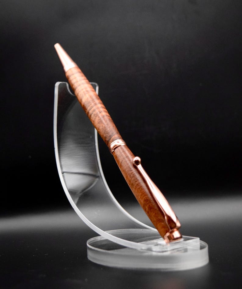 May include: A handcrafted wooden pen with a copper-colored clip and tip. The pen features a detailed wood grain pattern and is displayed on a clear acrylic stand against a dark background. The pen is a stylish writing instrument.