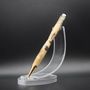 Puede incluir: Un bol&iacute;grafo artesanal con un cuerpo de veta de madera clara, acentuado con metal dorado e inclusiones de color p&uacute;rpura oscuro. El bol&iacute;grafo se exhibe en un soporte de acr&iacute;lico transparente, mostrando su elegante dise&ntilde;o. La punta del bol&iacute;grafo tambi&eacute;n es dorada.