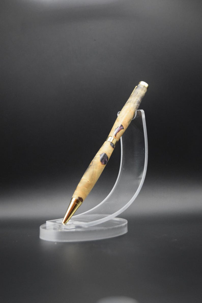 Puede incluir: Un bol&iacute;grafo artesanal con un cuerpo de veta de madera clara, acentuado con metal dorado e inclusiones de color p&uacute;rpura oscuro. El bol&iacute;grafo se exhibe en un soporte de acr&iacute;lico transparente, mostrando su elegante dise&ntilde;o. La punta del bol&iacute;grafo tambi&eacute;n es dorada.