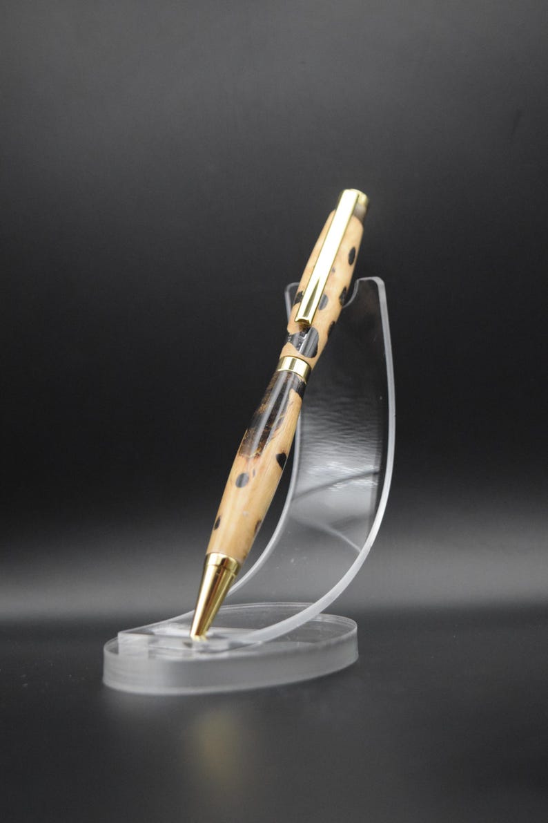 May include: A wooden pen with a black and brown patterned design and gold accents. The pen is displayed on a clear plastic stand.