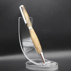 May include: A wooden pen with silver accents is displayed on a clear acrylic stand. The pen has a light brown wood grain pattern and a silver tip and clip. The stand is round at the base and has a curved support.