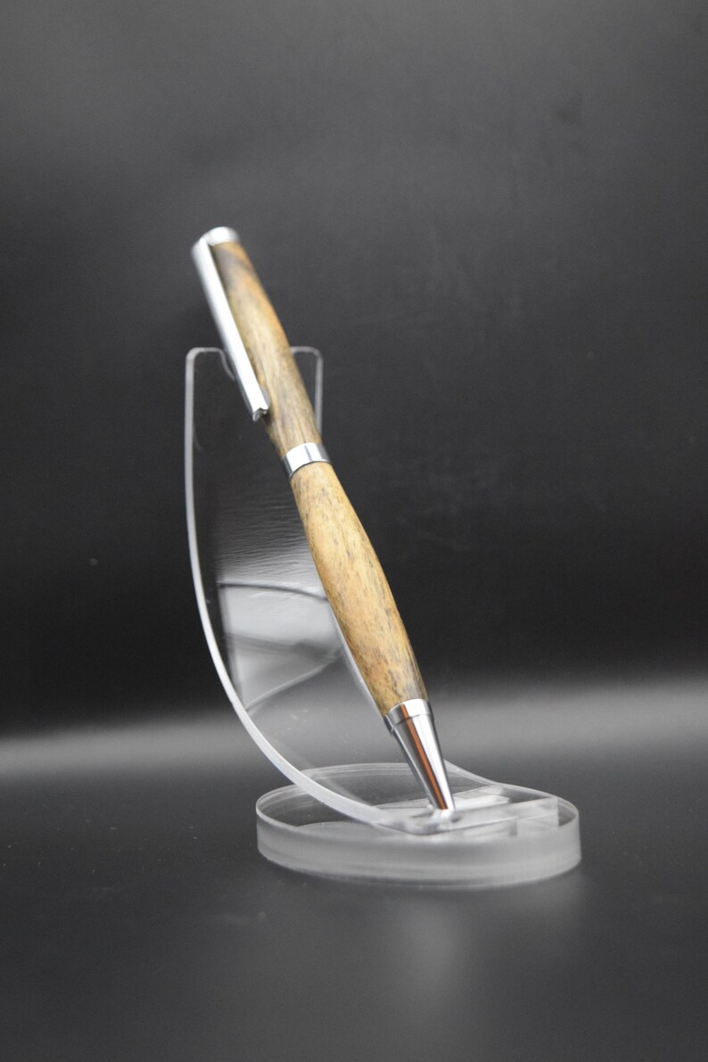 May include: A wooden pen with silver accents is displayed on a clear acrylic stand. The pen has a light brown wood grain pattern and a silver tip and clip. The stand is round at the base and has a curved support.