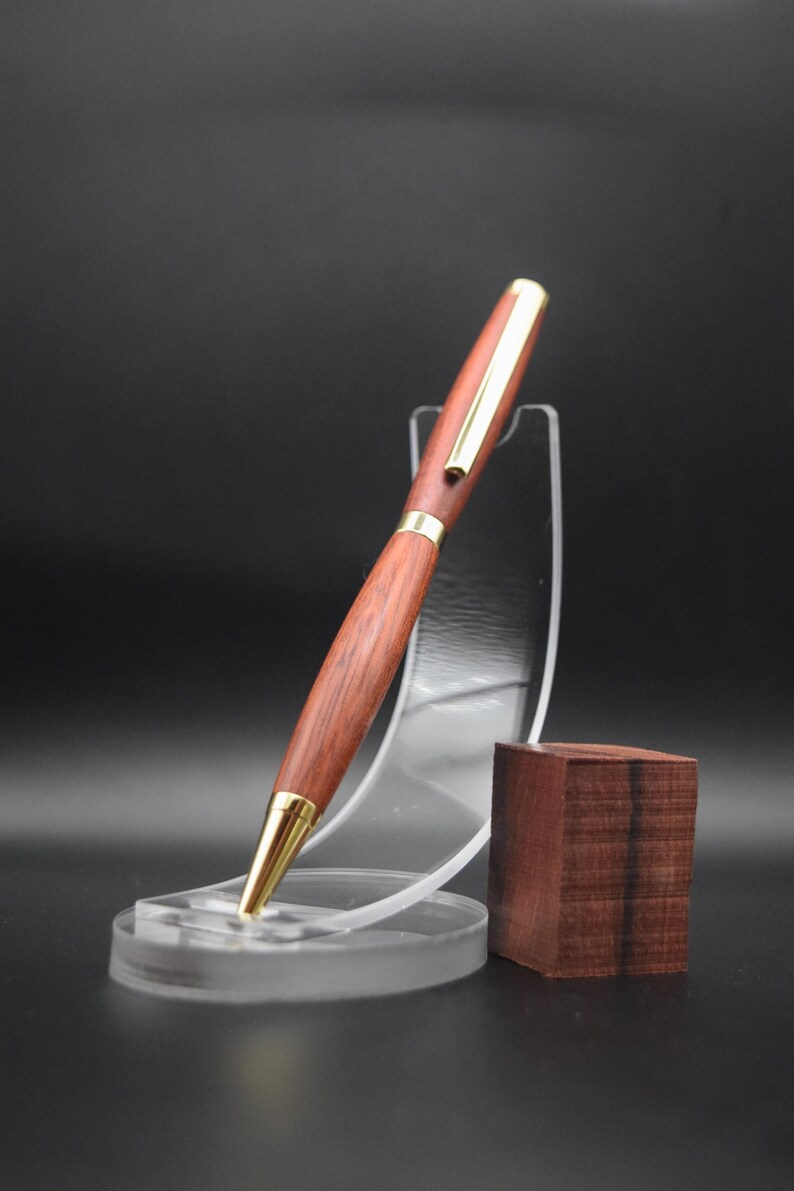 Puede incluir: Un bol&iacute;grafo de madera hecho a mano con un rico acabado marr&oacute;n rojizo y detalles dorados. El bol&iacute;grafo se exhibe sobre un soporte de acr&iacute;lico transparente, con un peque&ntilde;o bloque de madera al lado. El dise&ntilde;o del bol&iacute;grafo es elegante y cl&aacute;sico.