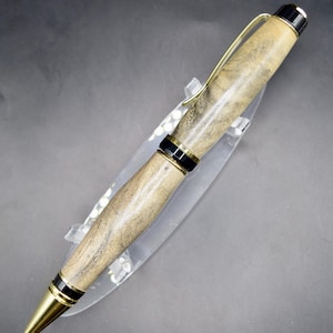 May include: A handcrafted pen with a light brown wooden barrel, accented with black and gold-colored metal details. The pen features a gold-colored clip and a gold-colored tip. The pen is displayed on a clear acrylic stand.