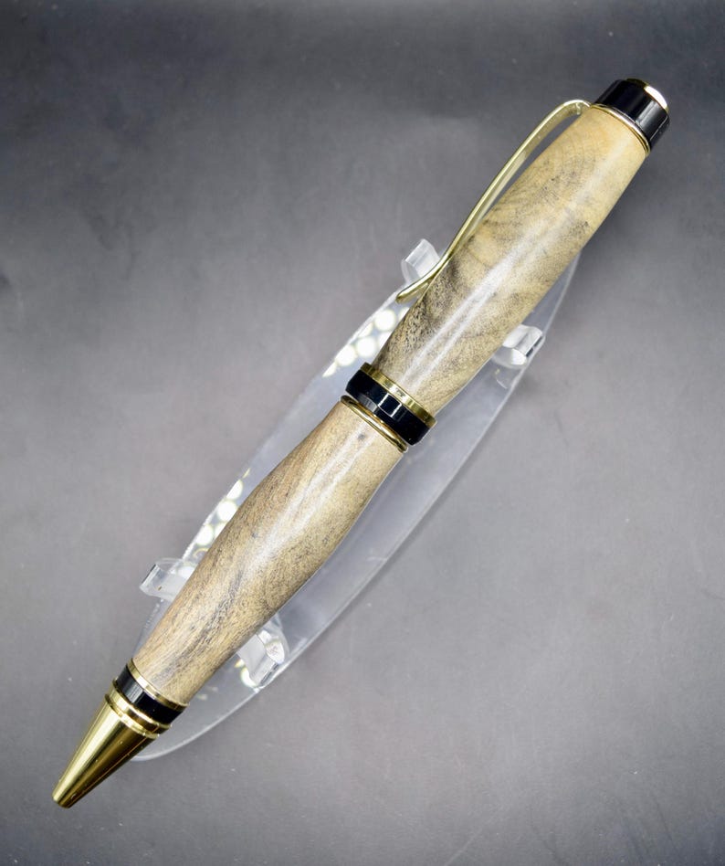 May include: A handcrafted pen with a light brown wooden barrel, accented with black and gold-colored metal details. The pen features a gold-colored clip and a gold-colored tip. The pen is displayed on a clear acrylic stand.