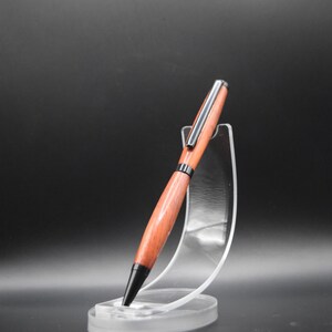 K&ouml;nnte beinhalten: Ein handgefertigter Holzstift mit rotbraunem Finish, akzentuiert durch silberne und schwarze Metallkomponenten. Der Stift wird auf einem transparenten Acrylst&auml;nder pr&auml;sentiert, der sein elegantes Design hervorhebt. Ein raffiniertes Schreibger&auml;t.