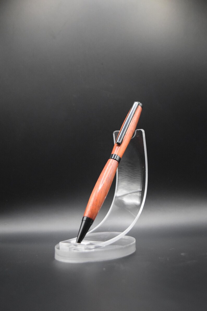 K&ouml;nnte beinhalten: Ein handgefertigter Holzstift mit rotbraunem Finish, akzentuiert durch silberne und schwarze Metallkomponenten. Der Stift wird auf einem transparenten Acrylst&auml;nder pr&auml;sentiert, der sein elegantes Design hervorhebt. Ein raffiniertes Schreibger&auml;t.