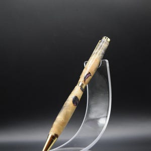 Puede incluir: Bol&iacute;grafo artesanal con un cuerpo de veta de madera clara, acentuado con inclusiones de color p&uacute;rpura oscuro. El bol&iacute;grafo presenta detalles met&aacute;licos dorados y una punta dorada. Se exhibe sobre un soporte de acr&iacute;lico transparente sobre un fondo oscuro.