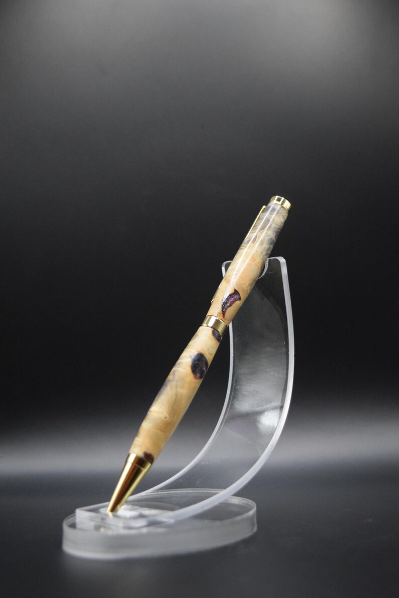 Puede incluir: Bol&iacute;grafo artesanal con un cuerpo de veta de madera clara, acentuado con inclusiones de color p&uacute;rpura oscuro. El bol&iacute;grafo presenta detalles met&aacute;licos dorados y una punta dorada. Se exhibe sobre un soporte de acr&iacute;lico transparente sobre un fondo oscuro.