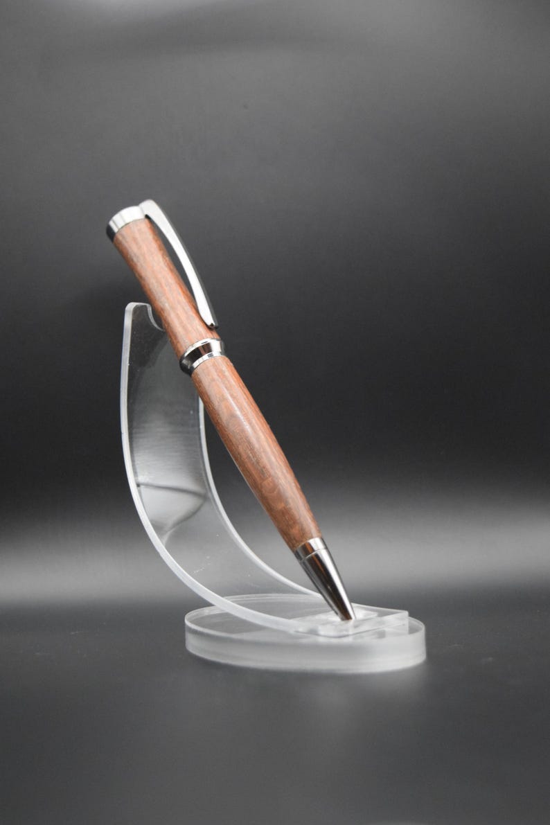 May include: A wooden pen with a silver trim, displayed on a clear plastic stand. The pen has a brown wood grain pattern.