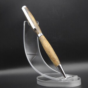 May include: A wooden pen with a silver clip and tip is displayed on a clear acrylic stand. The pen has a light brown wood grain pattern. The stand has a circular base and a curved support.