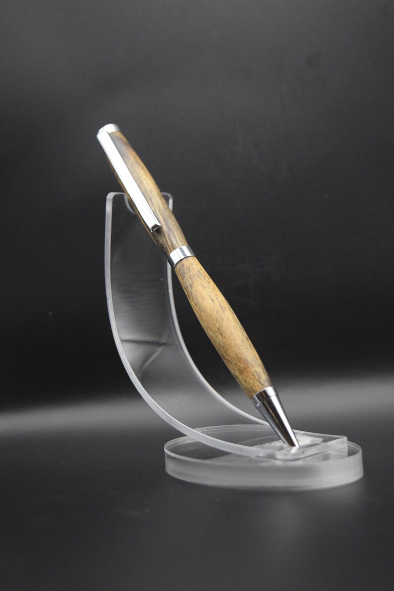 May include: A wooden pen with a silver clip and tip is displayed on a clear acrylic stand. The pen has a light brown wood grain pattern. The stand has a circular base and a curved support.