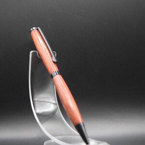 K&ouml;nnte beinhalten: Ein handgefertigter Holzstift mit rotbraunem Finish, schwarzen Akzenten und einem silbernen Clip. Der Stift wird auf einem transparenten Acrylst&auml;nder vor dunklem Hintergrund pr&auml;sentiert. Das Design des Stifts ist elegant.