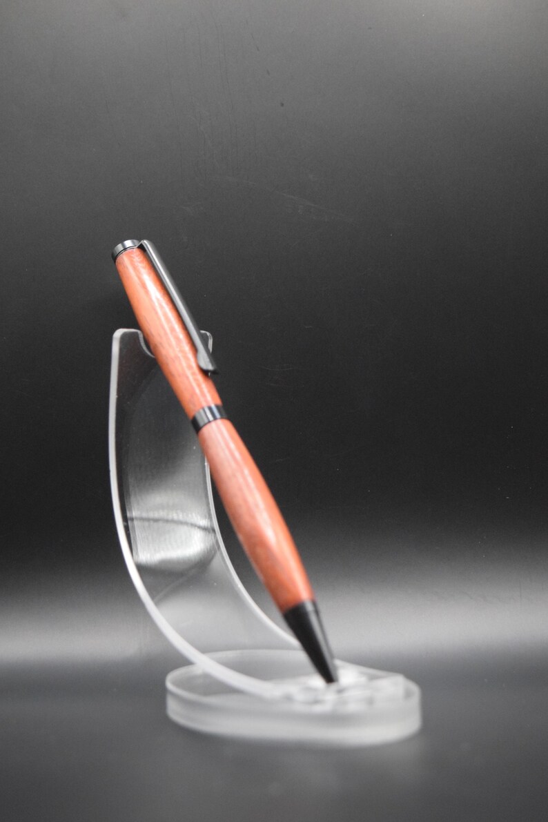 K&ouml;nnte beinhalten: Ein handgefertigter Holzstift mit rotbraunem Finish, schwarzen Akzenten und einem silbernen Clip. Der Stift wird auf einem transparenten Acrylst&auml;nder vor dunklem Hintergrund pr&auml;sentiert. Das Design des Stifts ist elegant.