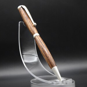 May include: A handcrafted wooden pen with a silver clip and tip, displayed on a clear acrylic stand. The pen features a rich, brown wood grain pattern and a sleek, modern design. The pen is approximately 6 inches long.