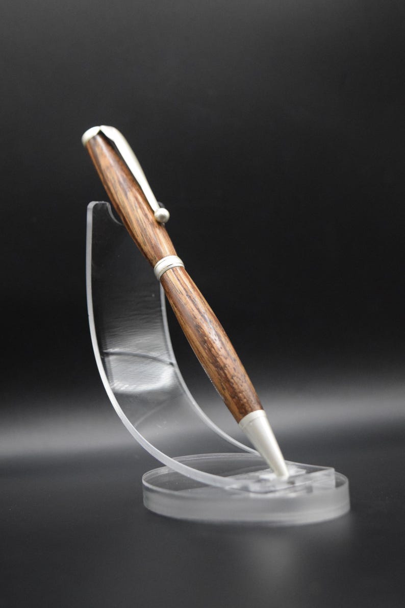 May include: A handcrafted wooden pen with a silver clip and tip, displayed on a clear acrylic stand. The pen features a rich, brown wood grain pattern and a sleek, modern design. The pen is approximately 6 inches long.