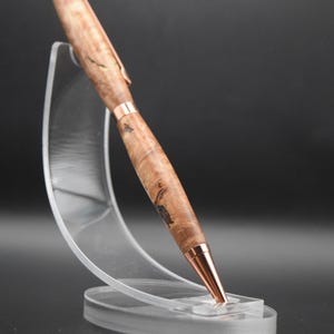 May include: A handcrafted wooden pen with a copper-colored clip and tip. The pen is made of light brown wood with dark brown and black patterns. The pen is displayed on a clear acrylic stand against a dark background.