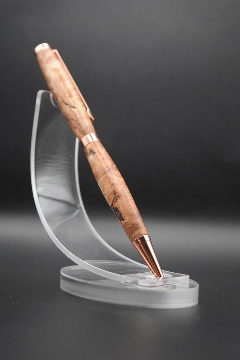 May include: A handcrafted wooden pen with a copper-colored clip and tip. The pen is made of light brown wood with dark brown and black patterns. The pen is displayed on a clear acrylic stand against a dark background.