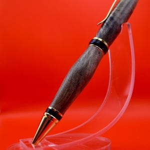 May include: A handcrafted wooden pen with a gold-colored clip and tip, accented with black and gold bands. The pen is displayed on a clear acrylic stand against a vibrant red background. The pen's body has a natural wood grain.