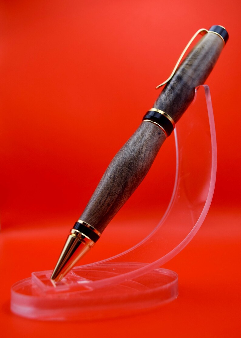 May include: A handcrafted wooden pen with a gold-colored clip and tip, accented with black and gold bands. The pen is displayed on a clear acrylic stand against a vibrant red background. The pen's body has a natural wood grain.