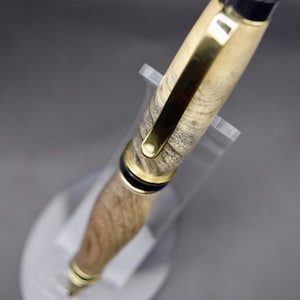 May include: A handcrafted wooden pen with gold-colored accents and a black cap. The pen features a natural wood grain pattern and is displayed on a clear acrylic stand. The pen's design includes a gold clip and a gold-colored tip.