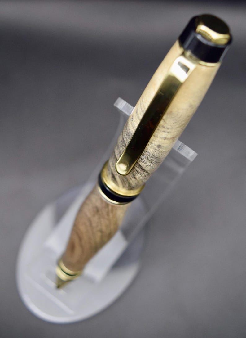 May include: A handcrafted wooden pen with gold-colored accents and a black cap. The pen features a natural wood grain pattern and is displayed on a clear acrylic stand. The pen's design includes a gold clip and a gold-colored tip.
