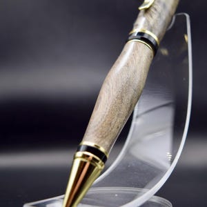 May include: A handcrafted wooden pen with a gold-colored clip and tip, accented with black bands. The pen has a natural wood grain finish and is displayed on a clear acrylic stand against a dark background.