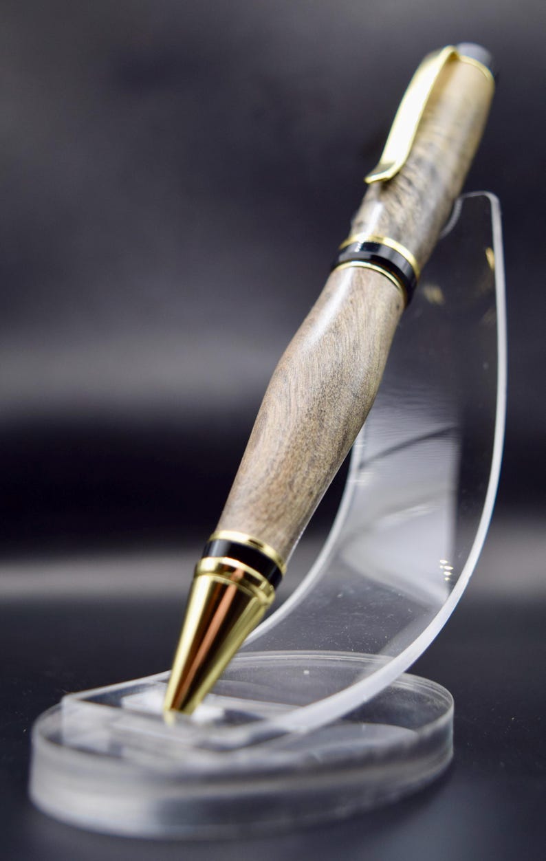 May include: A handcrafted wooden pen with a gold-colored clip and tip, accented with black bands. The pen has a natural wood grain finish and is displayed on a clear acrylic stand against a dark background.