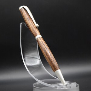 May include: A wooden pen with silver-colored accents is displayed on a clear acrylic stand. The pen has a brown, textured barrel and a silver clip. The pen's tip and the top are also silver. The stand is clear and circular.
