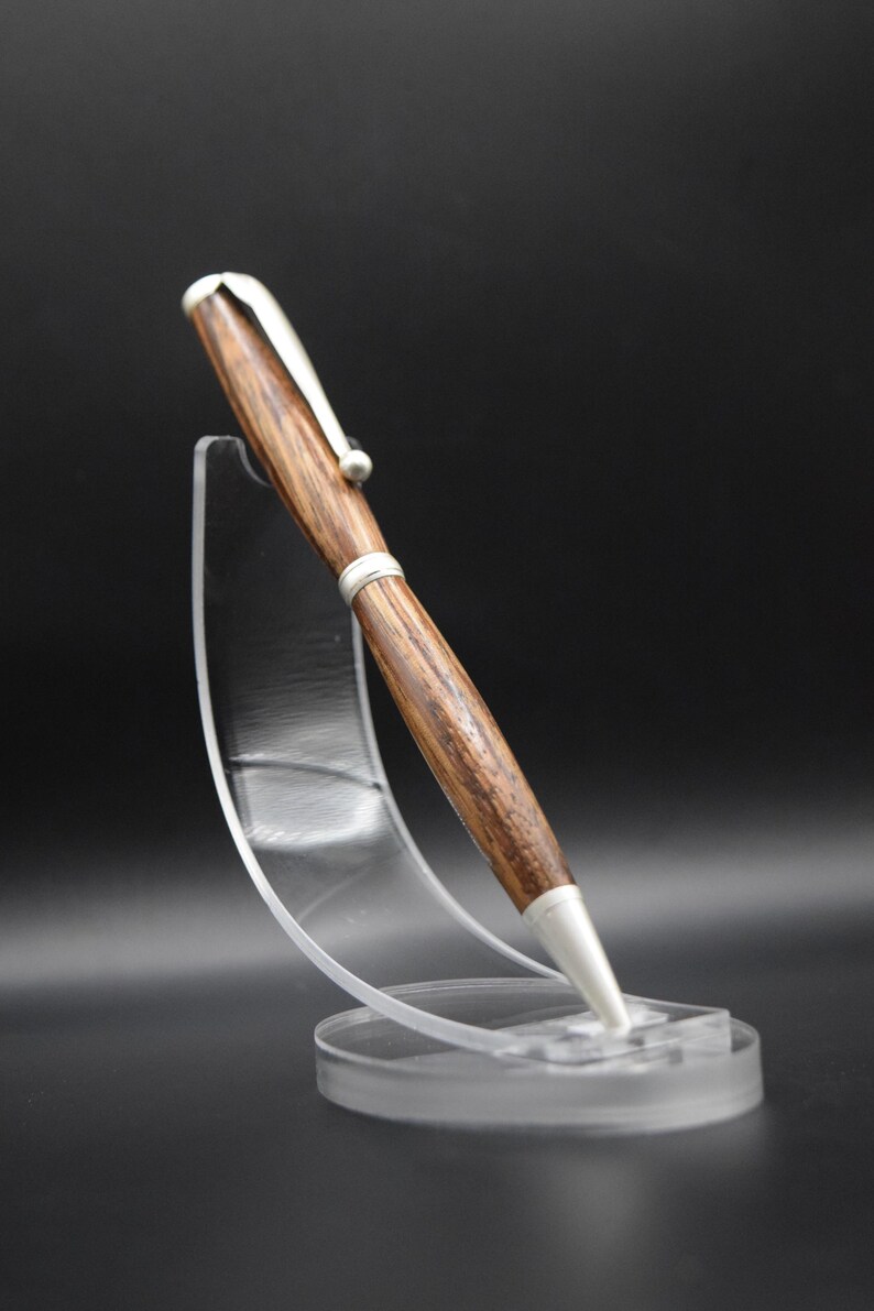May include: A wooden pen with silver-colored accents is displayed on a clear acrylic stand. The pen has a brown, textured barrel and a silver clip. The pen's tip and the top are also silver. The stand is clear and circular.