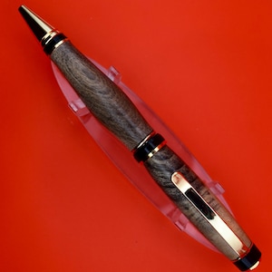May include: A handcrafted wooden pen with a gold-colored clip and accents. The pen features a dark wood grain pattern and a black tip. The pen is displayed on a clear plastic stand against a red background.