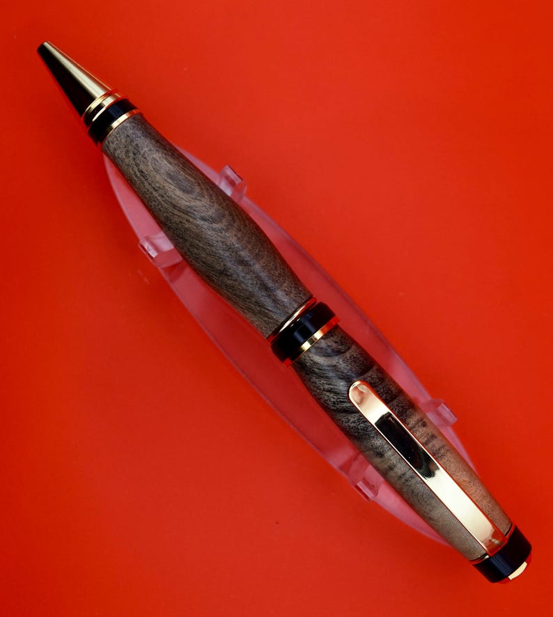 May include: A handcrafted wooden pen with a gold-colored clip and accents. The pen features a dark wood grain pattern and a black tip. The pen is displayed on a clear plastic stand against a red background.