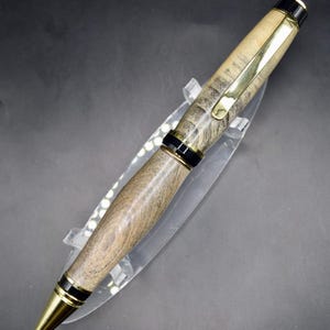May include: A handcrafted wooden pen with a gold-colored clip and tip. The pen features a light brown and gray wood grain pattern, accented by black bands. The pen is displayed on a clear plastic stand.