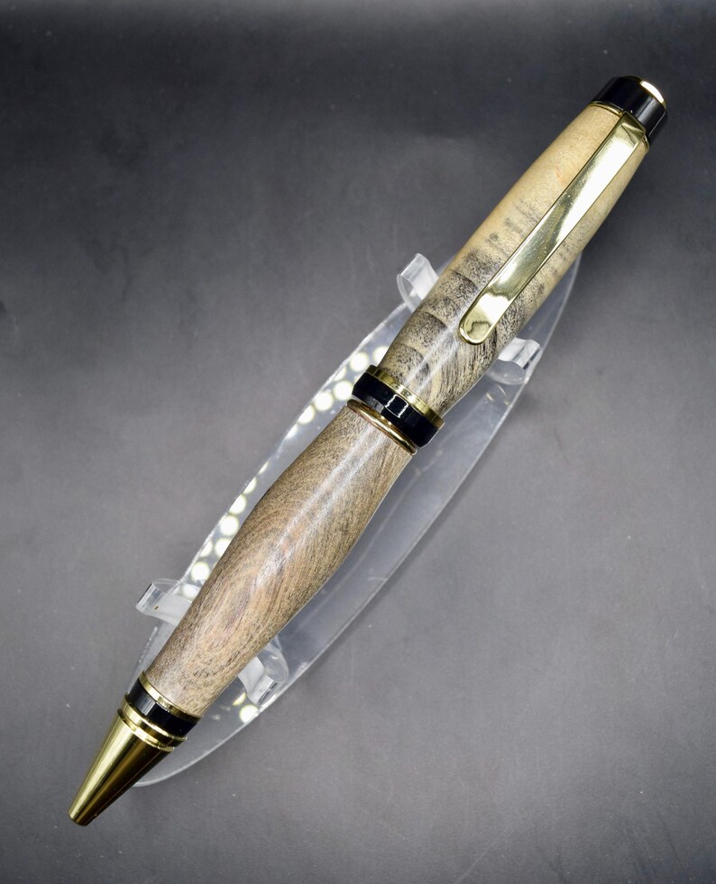 May include: A handcrafted wooden pen with a gold-colored clip and tip. The pen features a light brown and gray wood grain pattern, accented by black bands. The pen is displayed on a clear plastic stand.