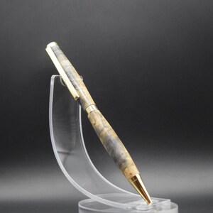 Puede incluir: Un bol&iacute;grafo artesanal con punta y clip dorados, y un cuerpo de madera veteada en tonos grises, beige y marr&oacute;n. El bol&iacute;grafo se exhibe sobre un soporte de acr&iacute;lico transparente sobre un fondo oscuro. Un elegante instrumento de escritura.