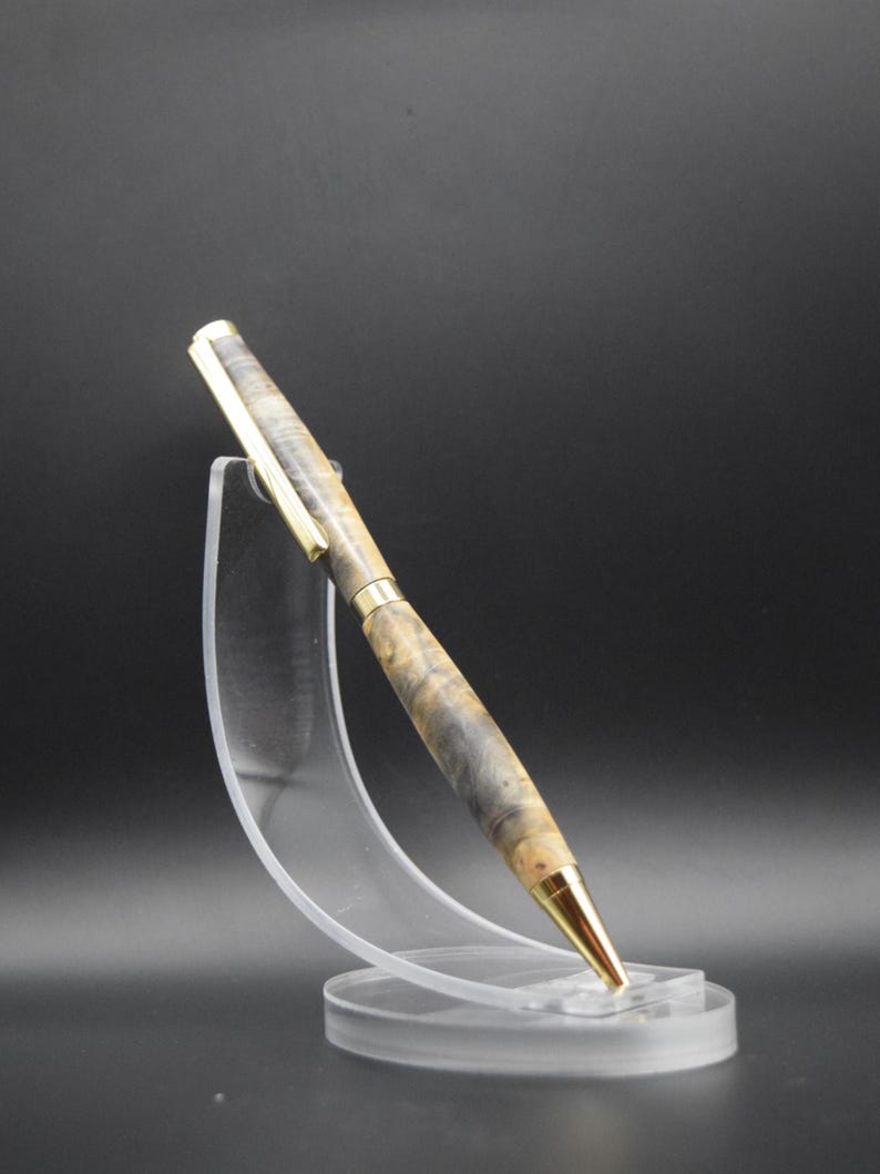 Puede incluir: Un bol&iacute;grafo artesanal con punta y clip dorados, y un cuerpo de madera veteada en tonos grises, beige y marr&oacute;n. El bol&iacute;grafo se exhibe sobre un soporte de acr&iacute;lico transparente sobre un fondo oscuro. Un elegante instrumento de escritura.