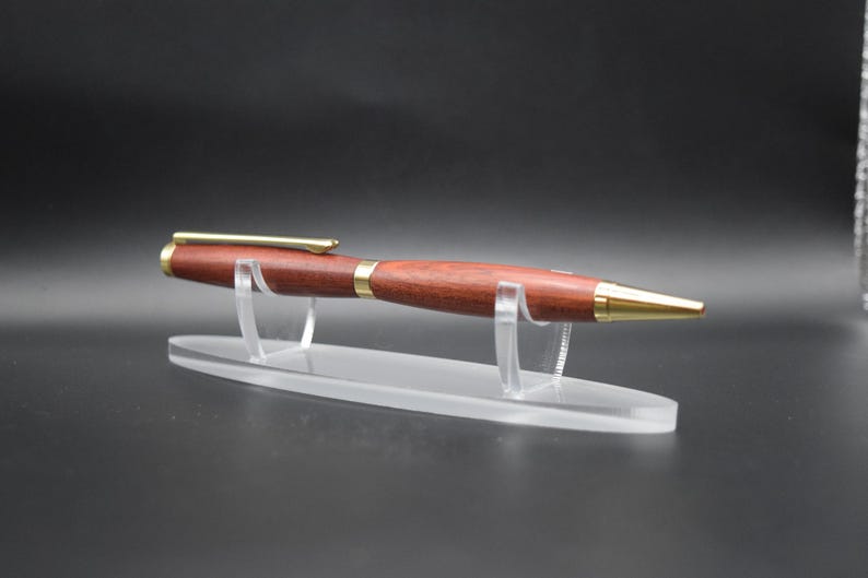 Puede incluir: Un bol&iacute;grafo de madera hecho a mano con un acabado marr&oacute;n rojizo y detalles dorados. El bol&iacute;grafo se exhibe en un soporte de acr&iacute;lico transparente, mostrando su elegante dise&ntilde;o. La punta y el clip del bol&iacute;grafo son dorados.