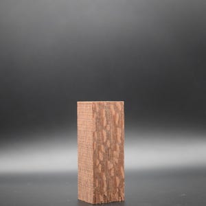 May include: A rectangular block of wood with a reddish-brown color and a unique, patterned grain. The wood block is sitting on a black surface.
