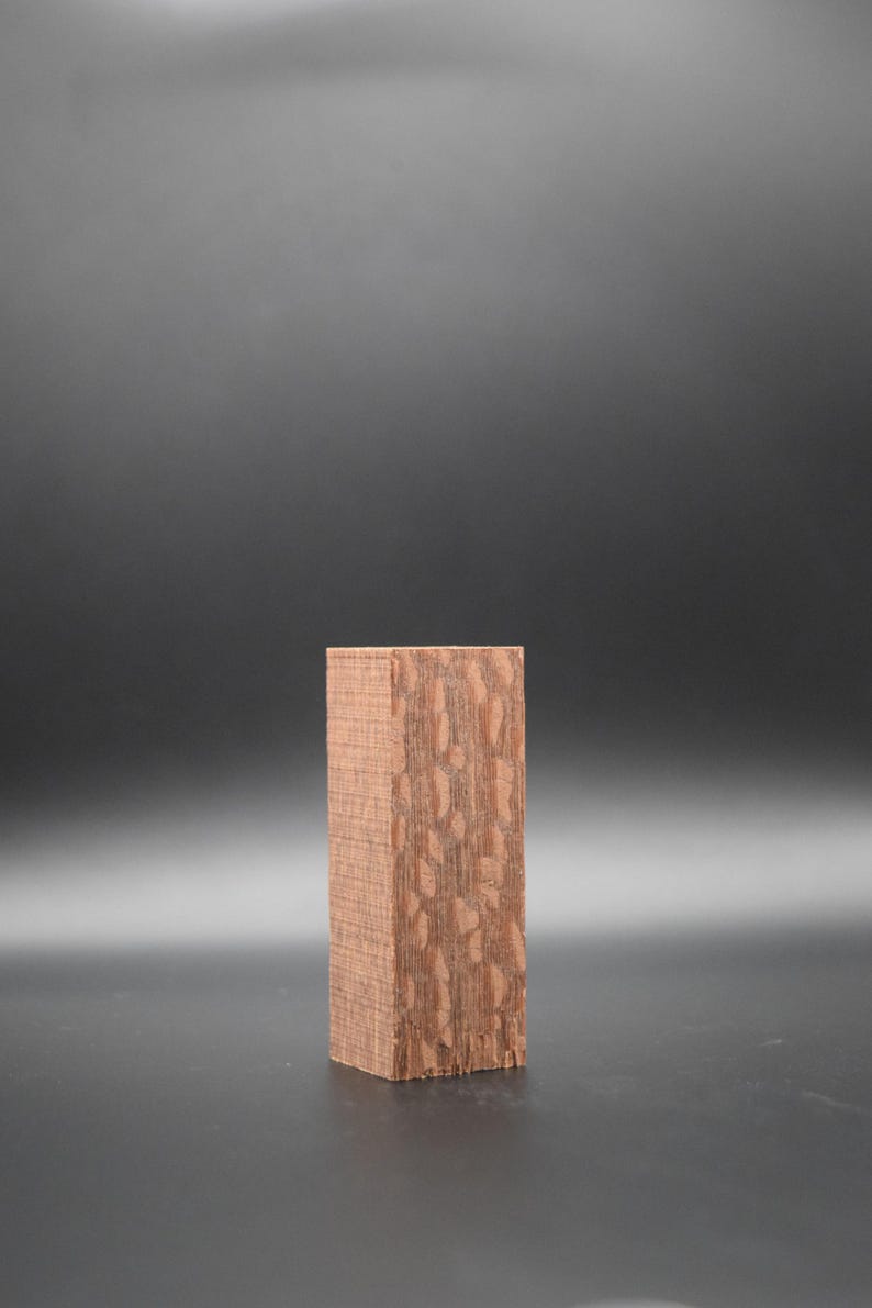 May include: A rectangular block of wood with a reddish-brown color and a unique, patterned grain. The wood block is sitting on a black surface.