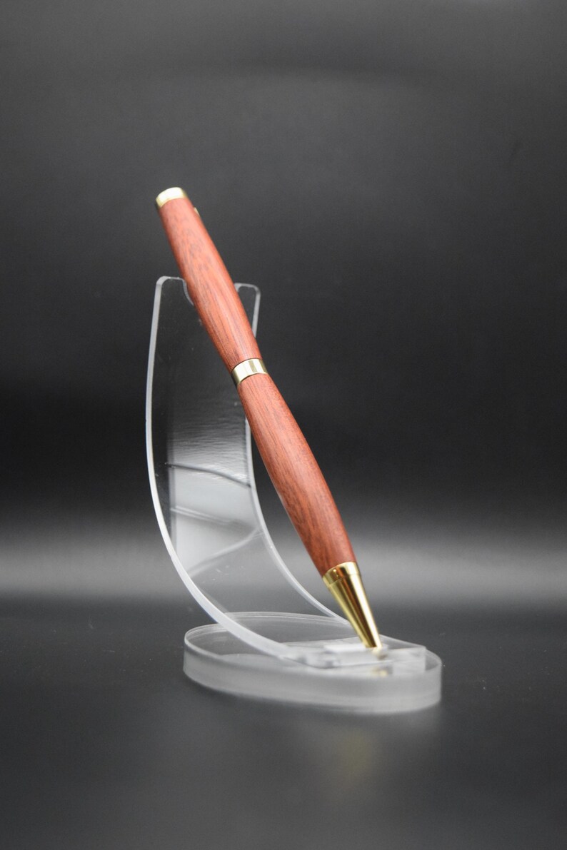 Puede incluir: Un bol&iacute;grafo de madera con un acabado marr&oacute;n intenso y detalles dorados. El bol&iacute;grafo se exhibe en un soporte de acr&iacute;lico transparente, mostrando su elegante dise&ntilde;o. La punta y la banda central del bol&iacute;grafo son doradas.