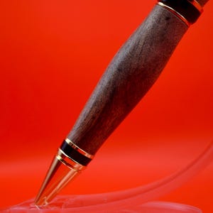 May include: A handcrafted wooden pen with a dark brown finish, accented by gold-colored metal bands and a gold-colored tip. The pen is displayed against a vibrant red background.